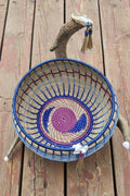 A handwoven basket with a blue rim and a purple and blue spiral pattern inside, displayed on a rustic wooden surface with a decorative branch prop.