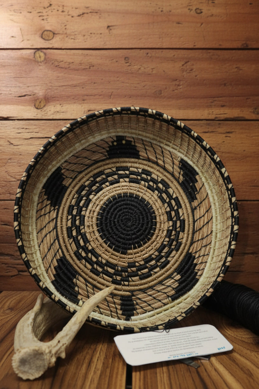 A hand-woven basket with a black and beige geometric pattern, displayed on a rustic wooden surface next to a small antler and a product tag.