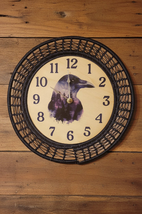 A round wall clock with a dark woven basket-style frame, black numerals, and a central illustration of a raven over a forest. The clock is mounted on a light gray wall above a wooden surface with small hanging items.
